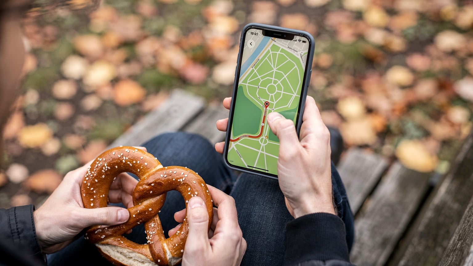Ein Reisender zeigt sein Smartphone-Display, auf dem eine Offline-Karte von Berlin mit einem markierten Wanderweg in einem Park zu sehen ist, während die Hände eines anderen Reisenden ein traditionelles deutsches Laugengebäck halten.
