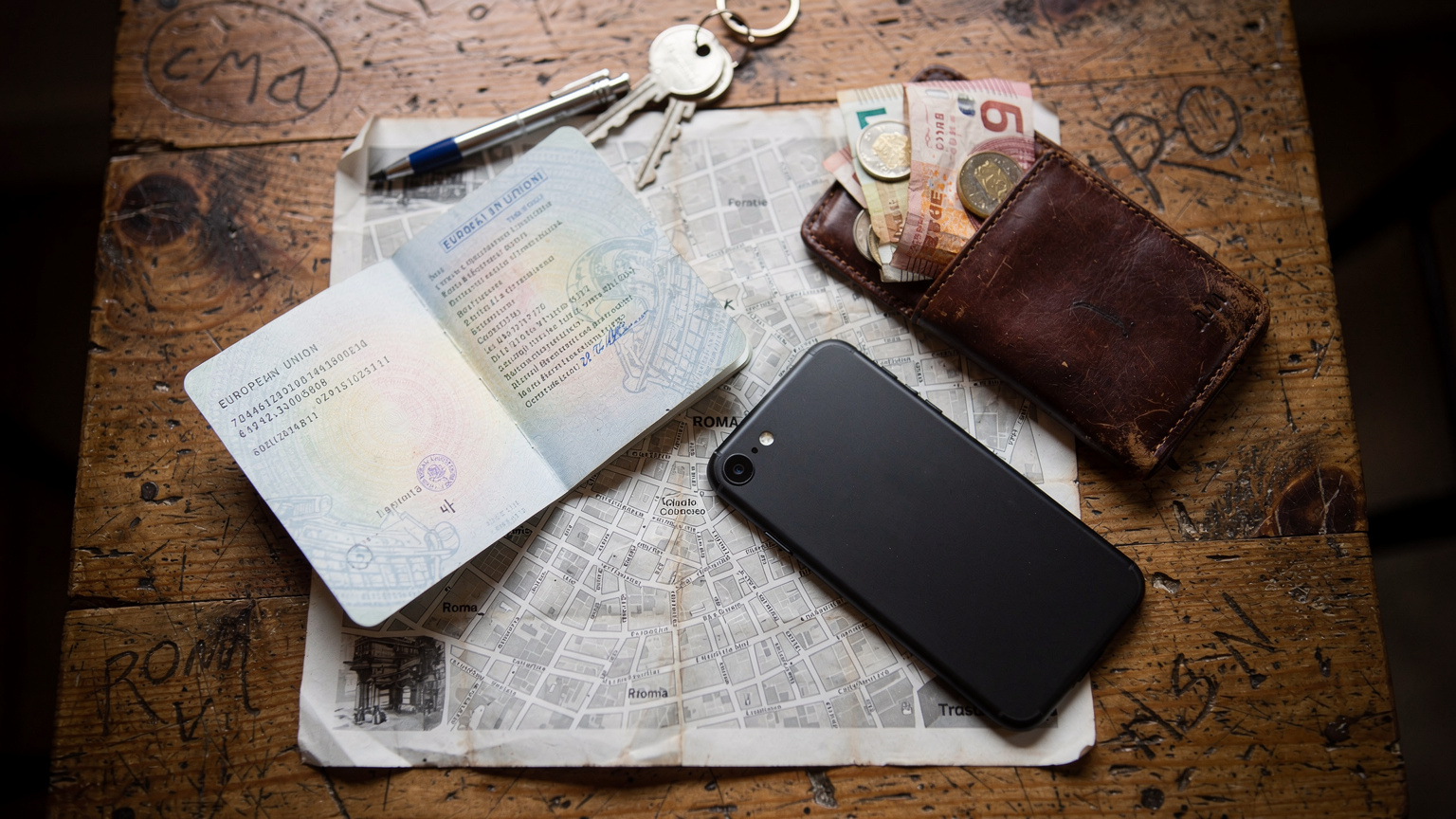 An overhead flat-lay of travel essentials: a passport, a smartphone, a small local currency wallet, and a folded paper map, all laid out on a worn wooden hostel table.
