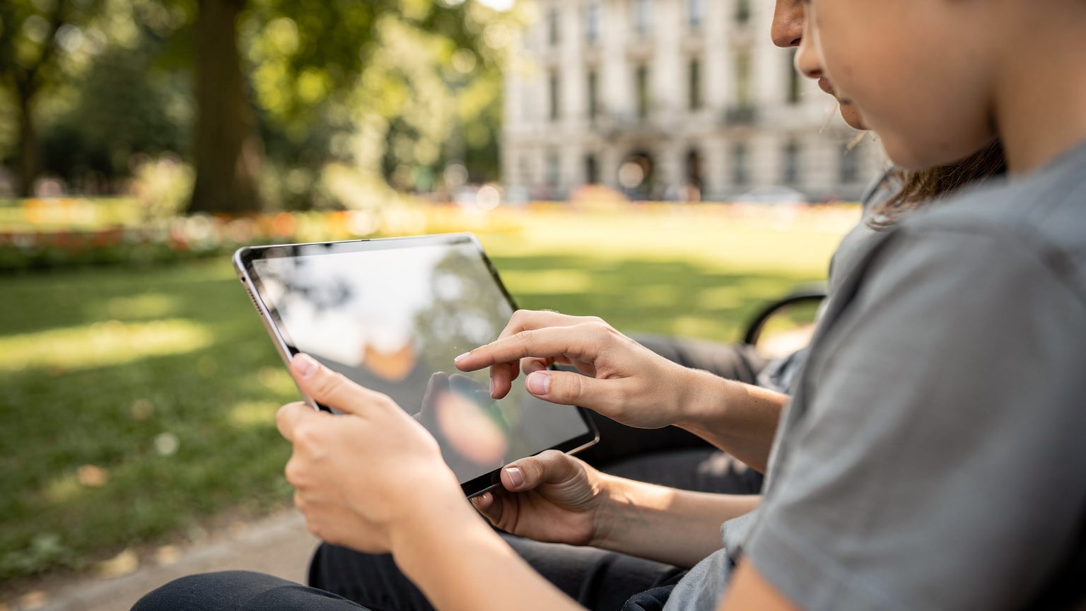Mani di una madre che aiuta il figlio di 10 anni a configurare un tablet con un'app di parental control mentre sono seduti su una panchina in un parco a Bruxelles, Belgio.