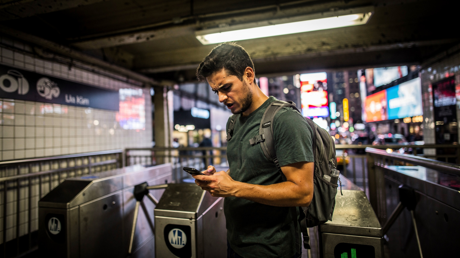 A person standing at a transit ticket gate for the MTA in New York City, looking at their phone with a frustrated expression, with the vibrant, blurry lights of Times Square visible in the background.