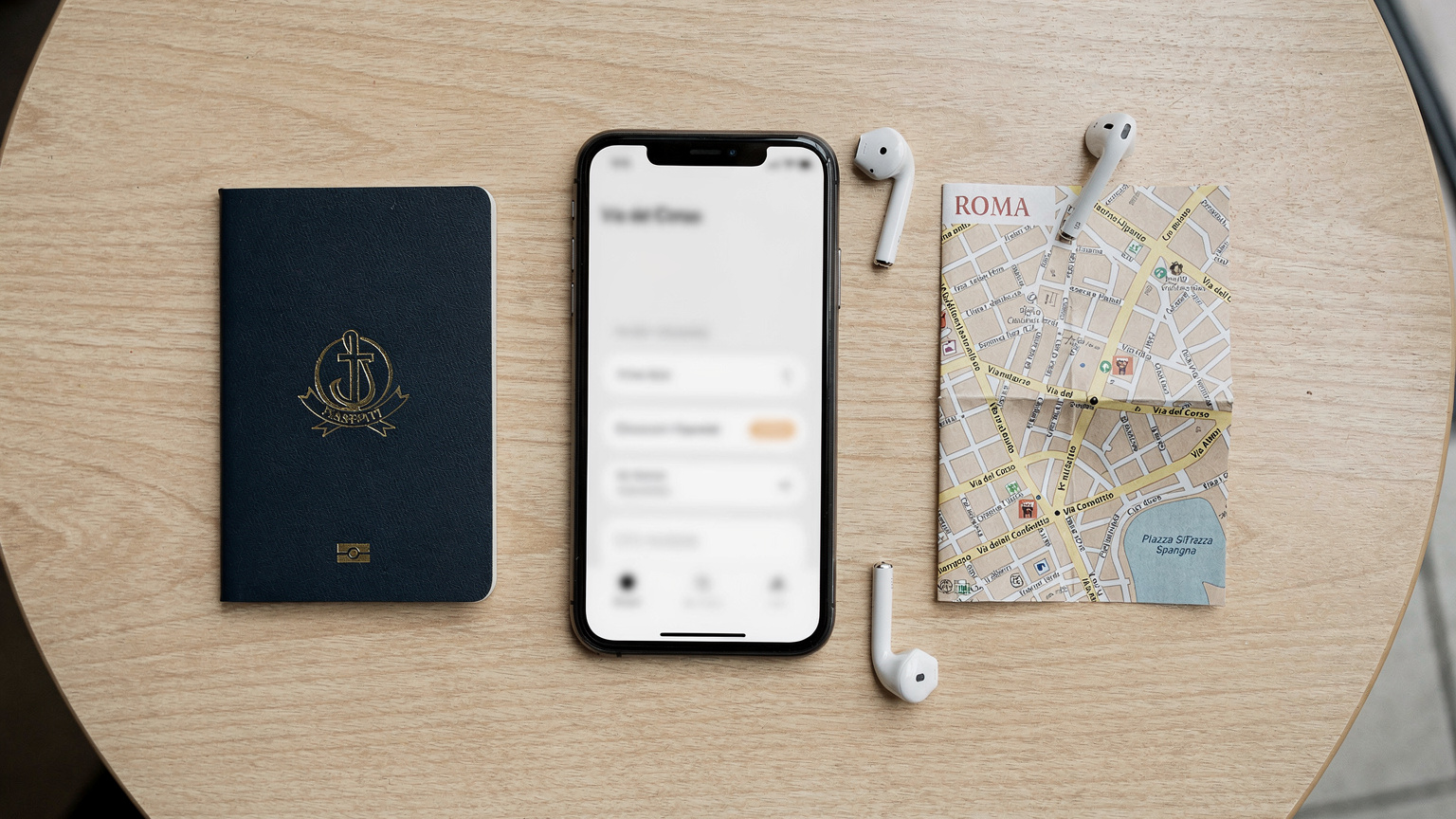 An overhead flat-lay shot of a passport, a smartphone displaying a settings menu (no readable text), a pair of wireless earbuds, and a small, folded map of Rome, Italy, on a light-colored cafe table.