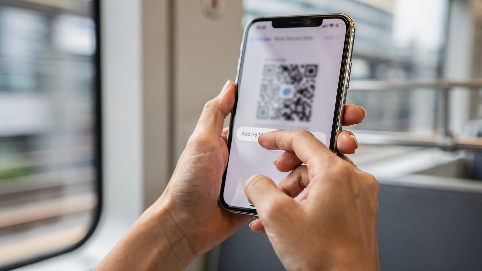 一位亞洲年輕女性的手在iPhone上操作eSIM設定介面,螢幕上模糊顯示著QR碼掃描的提示,背景是香港地鐵車廂的內部。