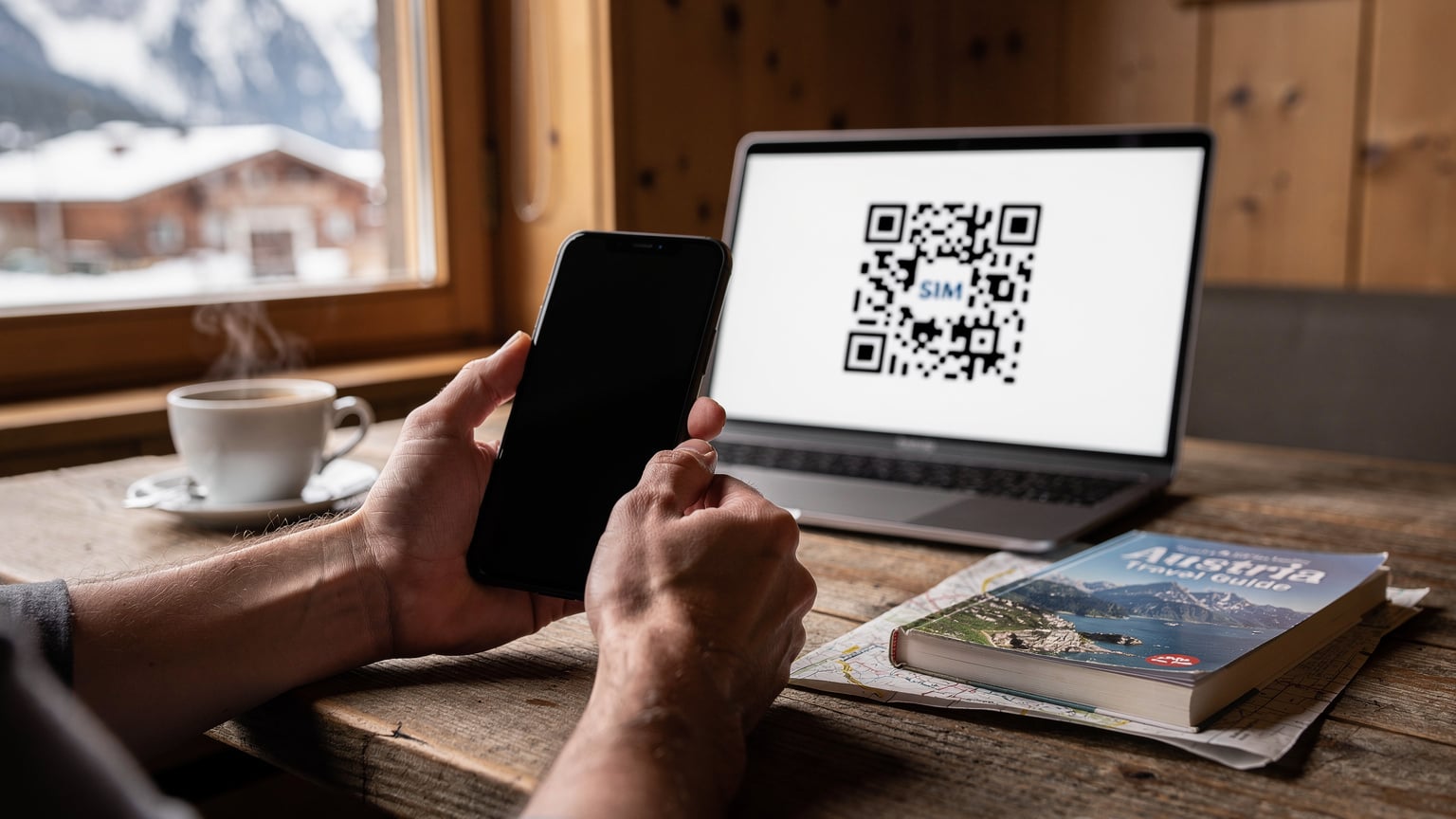 En detaljert nærbilde av hender som holder en smarttelefon, med en eSIM-installasjons-QR-kode synlig på en dataskjerm i bakgrunnen. En kopp kaffe og en reisebok ligger på et kafébord i en alpelandsby.