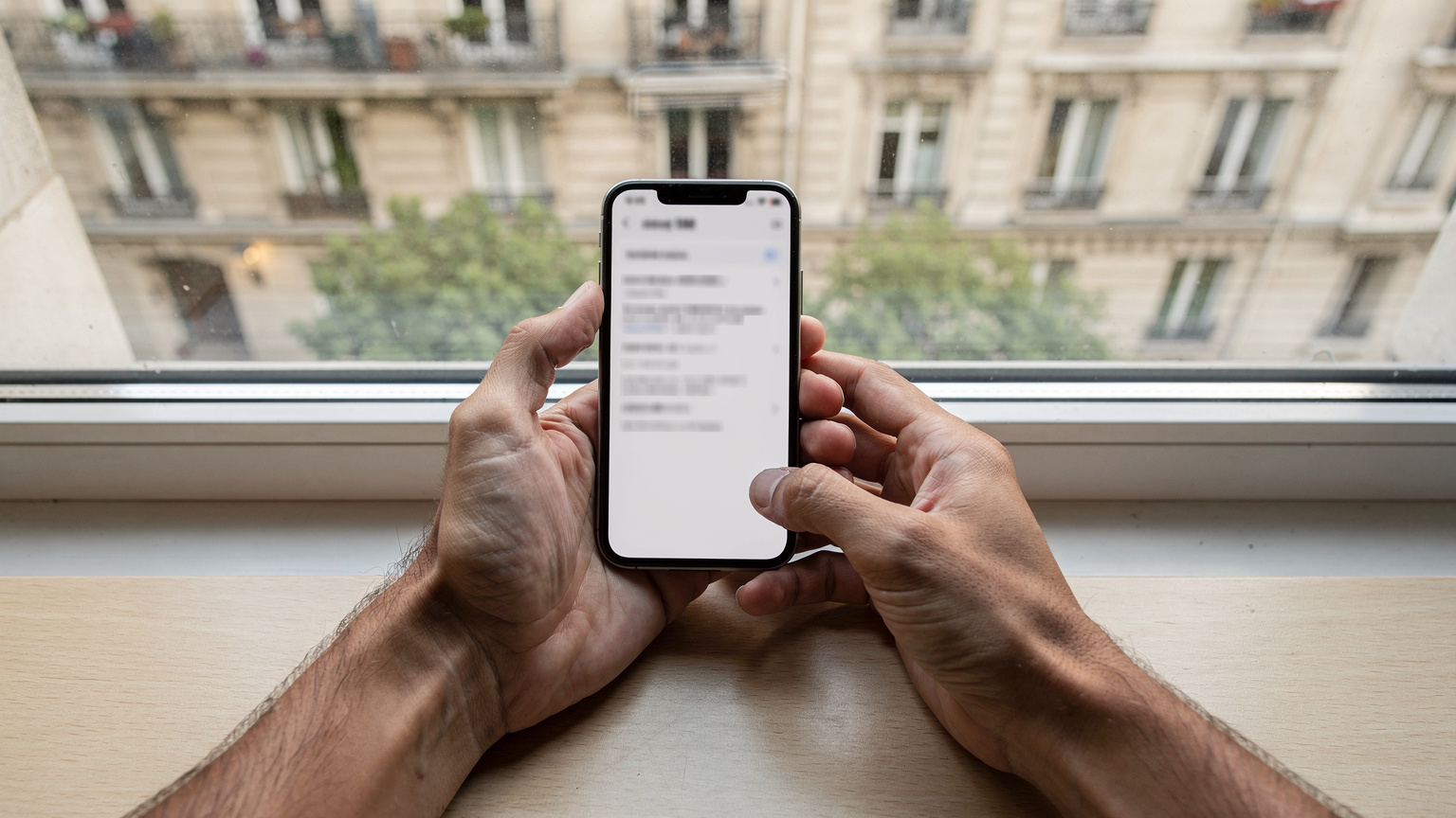 Ένα ζευγάρι χέρια κρατούν ένα smartphone, δείχνοντας στην οθόνη την επιλογή προσθήκης eSIM στις ρυθμίσεις του κινητού, με φόντο ένα παράθυρο που βλέπει σε μια τυπική παριζιάνικη λεωφόρο.
