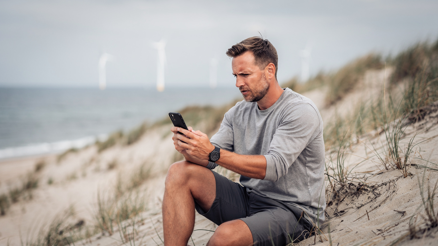 Mężczyzna siedzący na wydmie, przeglądający mapę na smartfonie, z wiatrakami w tle na horyzoncie niemieckiego wybrzeża Bałtyku.