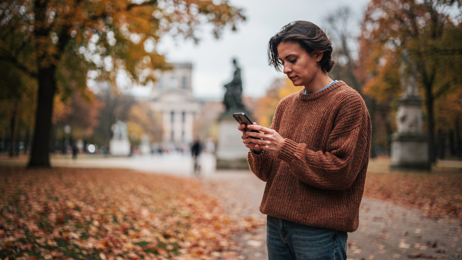 A traveler in Berlin's Tiergarten during autumn, checking their phone for directions with fallen leaves around.