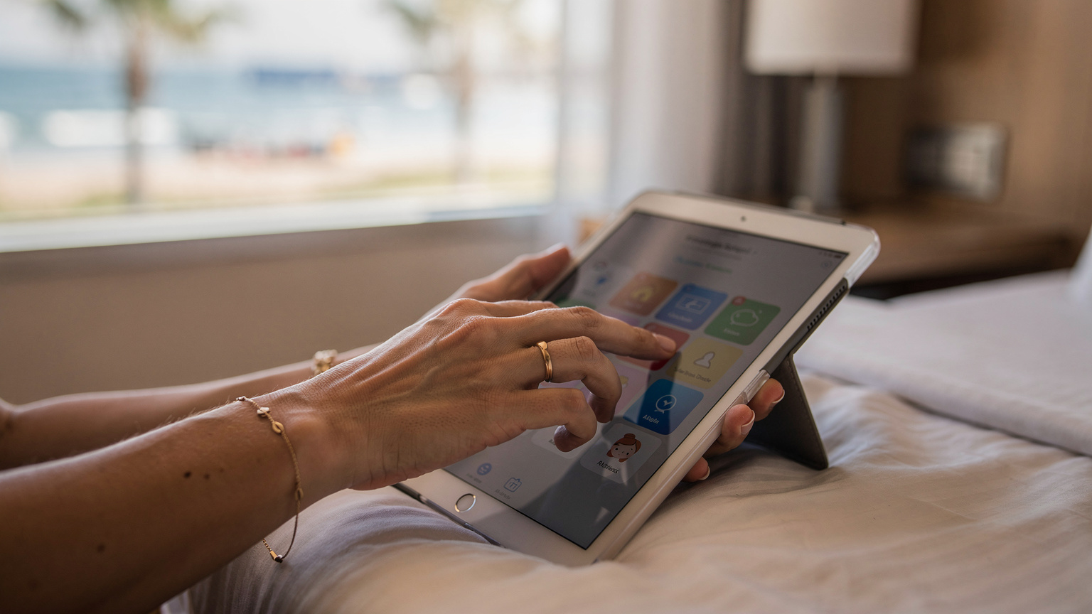 O mână de părinte, o femeie, configurează setările de timp de ecran și control parental pe un iPad pentru copii, într-o cameră de hotel modernă din Antalya, Turcia. Lumina caldă a dimineții pătrunde pe fereastră, iar în fundal se zărește o plajă. Gestul indică control și planificare.