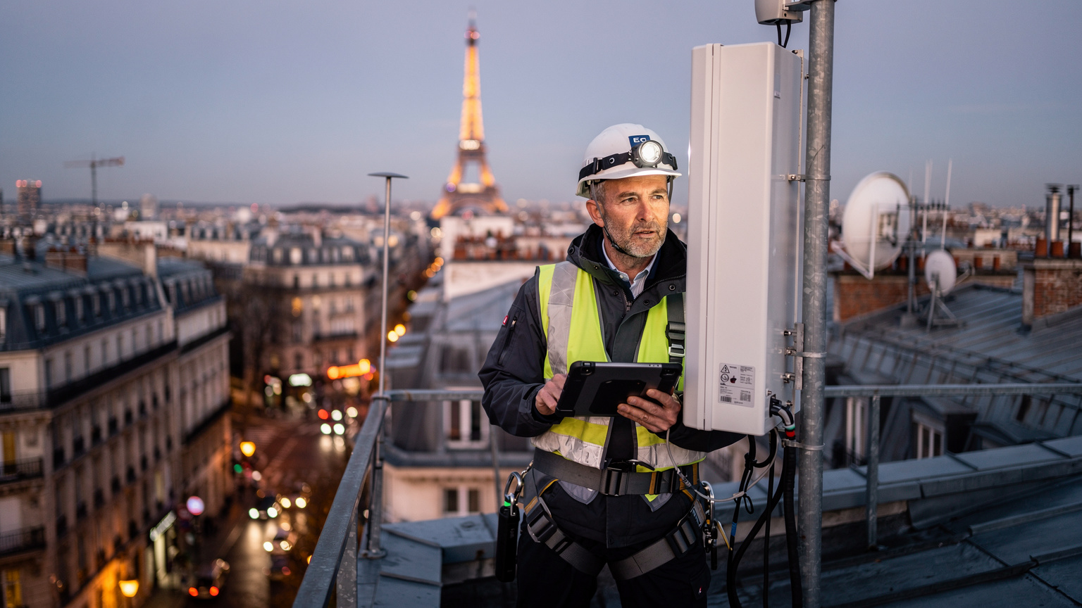 Een techneut inspecteert een 5G-antenne op een Parijs dak, met de stad als achtergrond, om de gereedheid voor de Olympische Spelen te waarborgen.