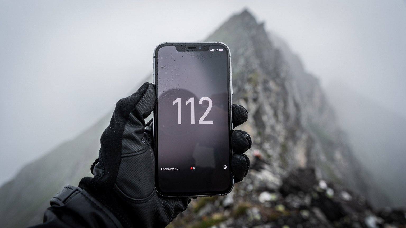 Tangan seorang pendaki sedang memegang smartphone dengan layar darurat menunjukkan nomor kontak SOS Eropa (112), di latar belakang terlihat kabut tebal menyelimuti puncak gunung, memberikan kesan mendesak.