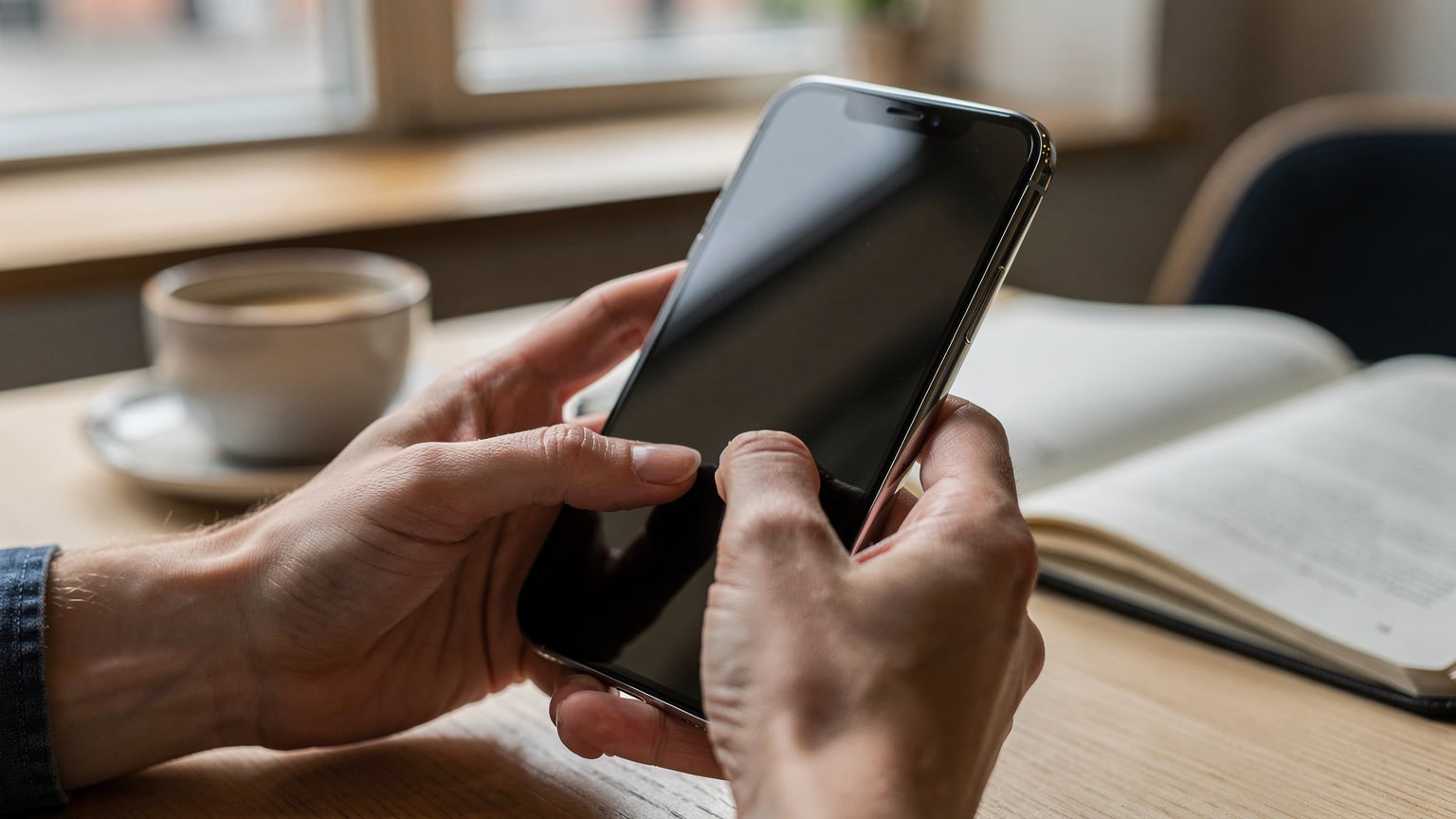 Bir kafede kahvesini yudumlarken, yeni iPhone 18'ini elinde tutan ve eSIM ayarlarını inceleyen bir gezginin elleri ve telefonun yakın çekimi.