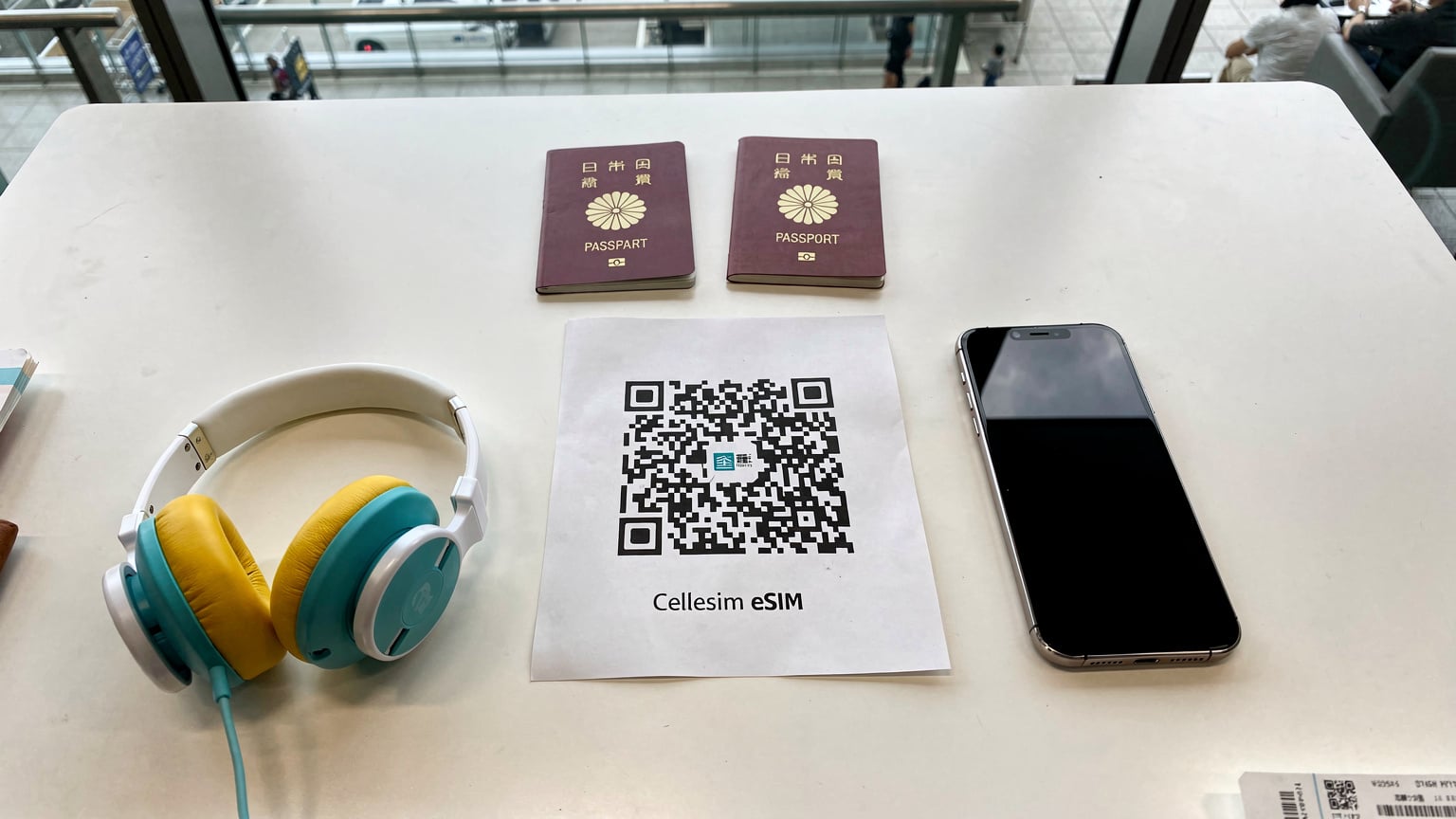 成田空港の出発ロビーで、搭乗前の最終確認をする家族の俯瞰ショット。パスポート、CellesimのeSIM情報が印刷されたQRコード、スマホ、そして子供のヘッドフォンがテーブルに並べられている。