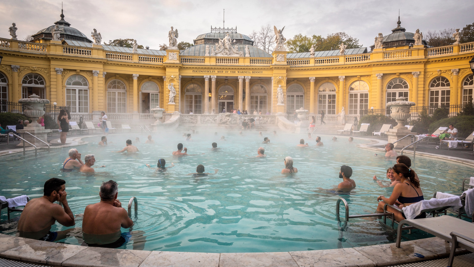Tourists and locals alike enjoy the warm thermal waters of the Széchenyi Thermal Bath in Budapest, steam rising into the cool air, under a grand architectural backdrop.