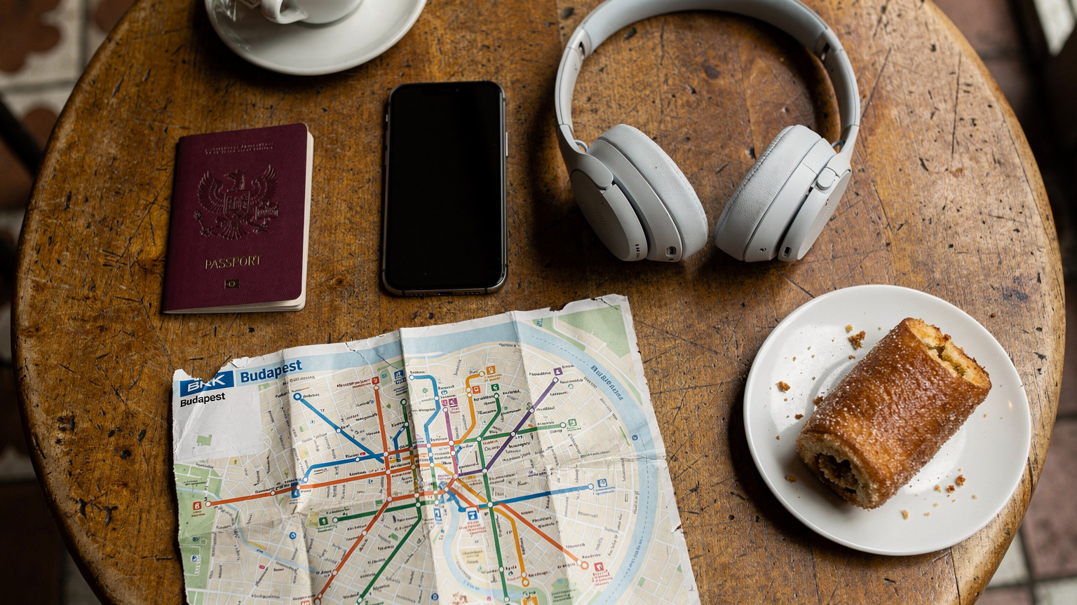 An overhead flat-lay of travel essentials for Hungary, including a passport, a smartphone with a Cellesim logo (blurred), noise-canceling headphones, a local Budapest transit map, and a traditional Hungarian pastry.
