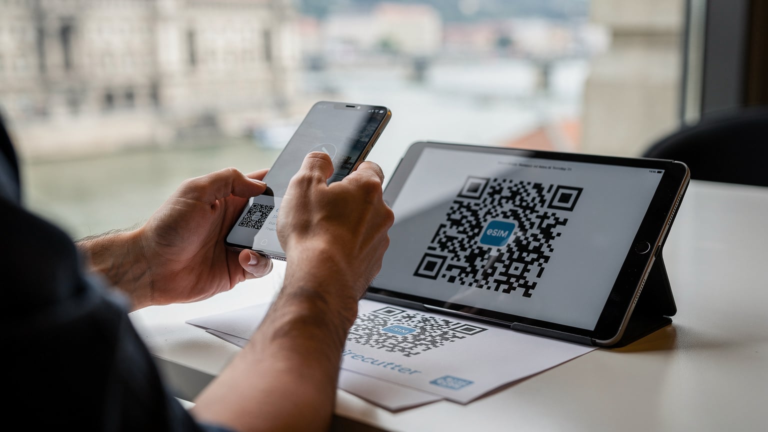 A close-up shot of human hands holding a smartphone, with one hand pointing at an eSIM QR code displayed on another device, illustrating the ease of digital activation.