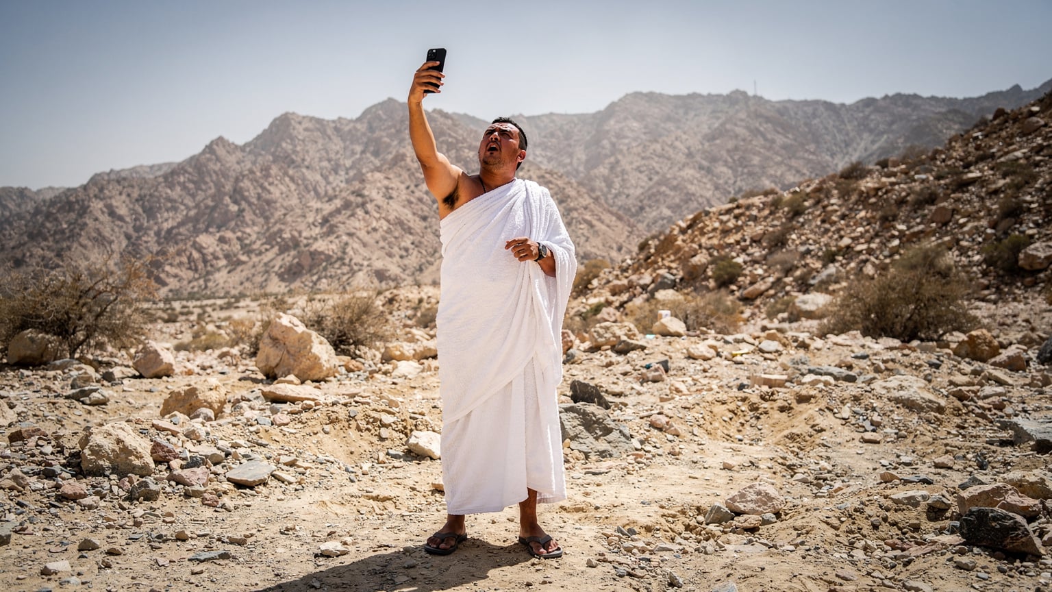 Seorang jemaah haji sedang cuba mendapatkan isyarat telefon di kawasan pergunungan di luar Makkah, menunjukkan cabaran sambungan di kawasan pedalaman.