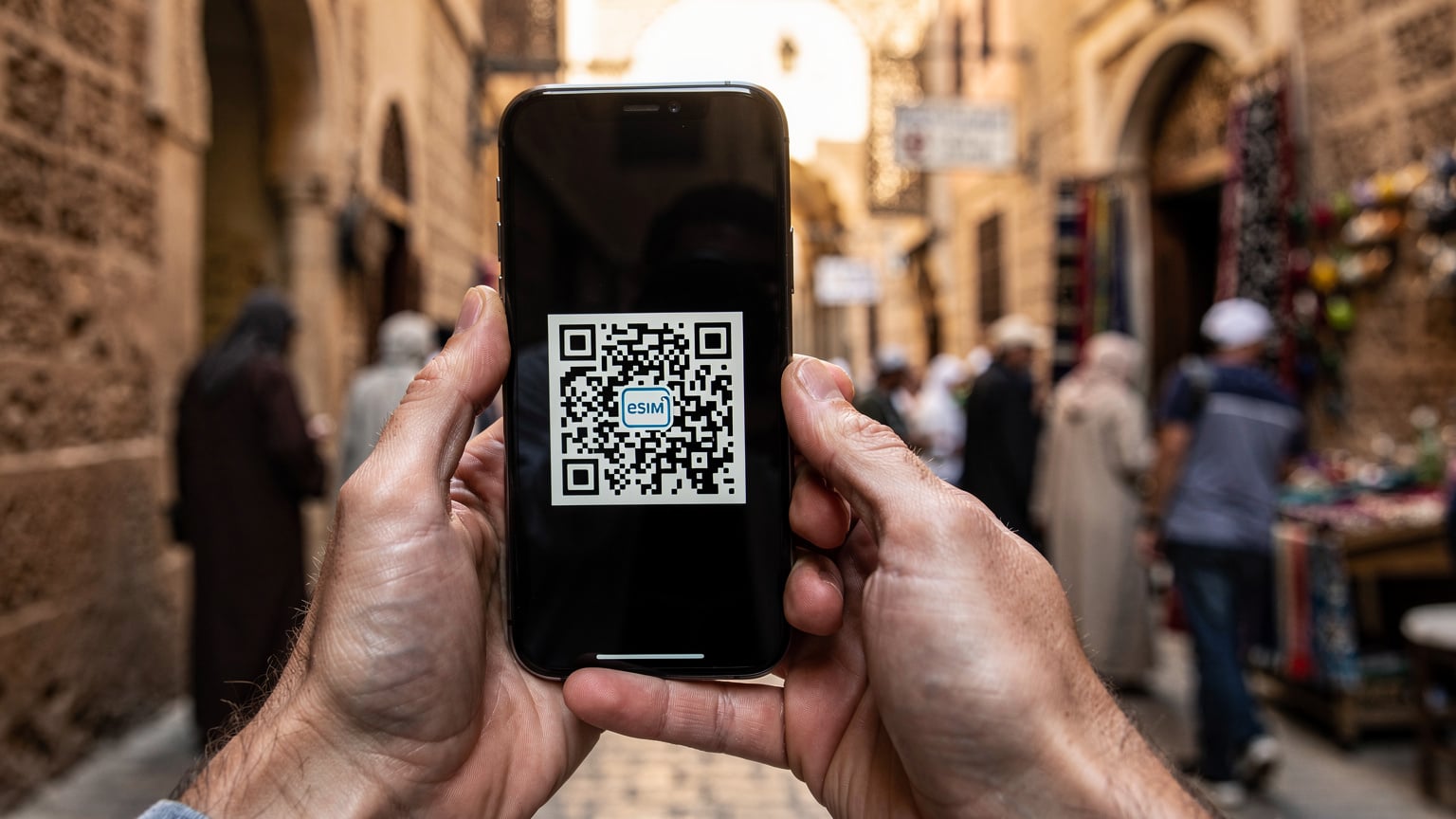 Zbliżenie na dłonie trzymające smartfon, na ekranie widać proces instalacji eSIM z kodem QR, w tle rozmazane uliczki medyny.