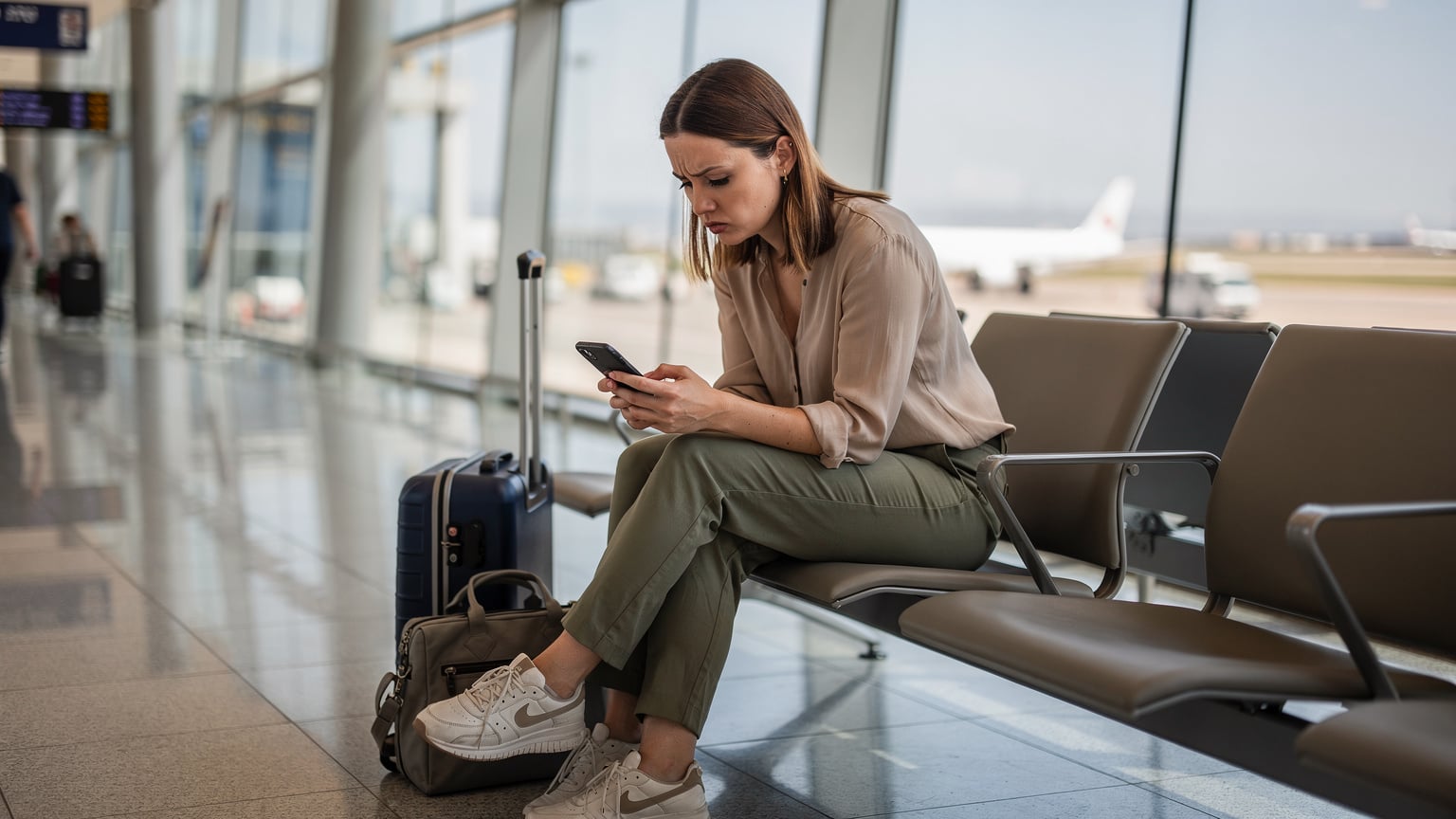 Młoda kobieta siedząca na ławce na lotnisku w Marrakeszu, z zaniepokojeniem patrzy na swój telefon, obok niej bagaż podręczny, symbolizujący problem z brakiem łączności.