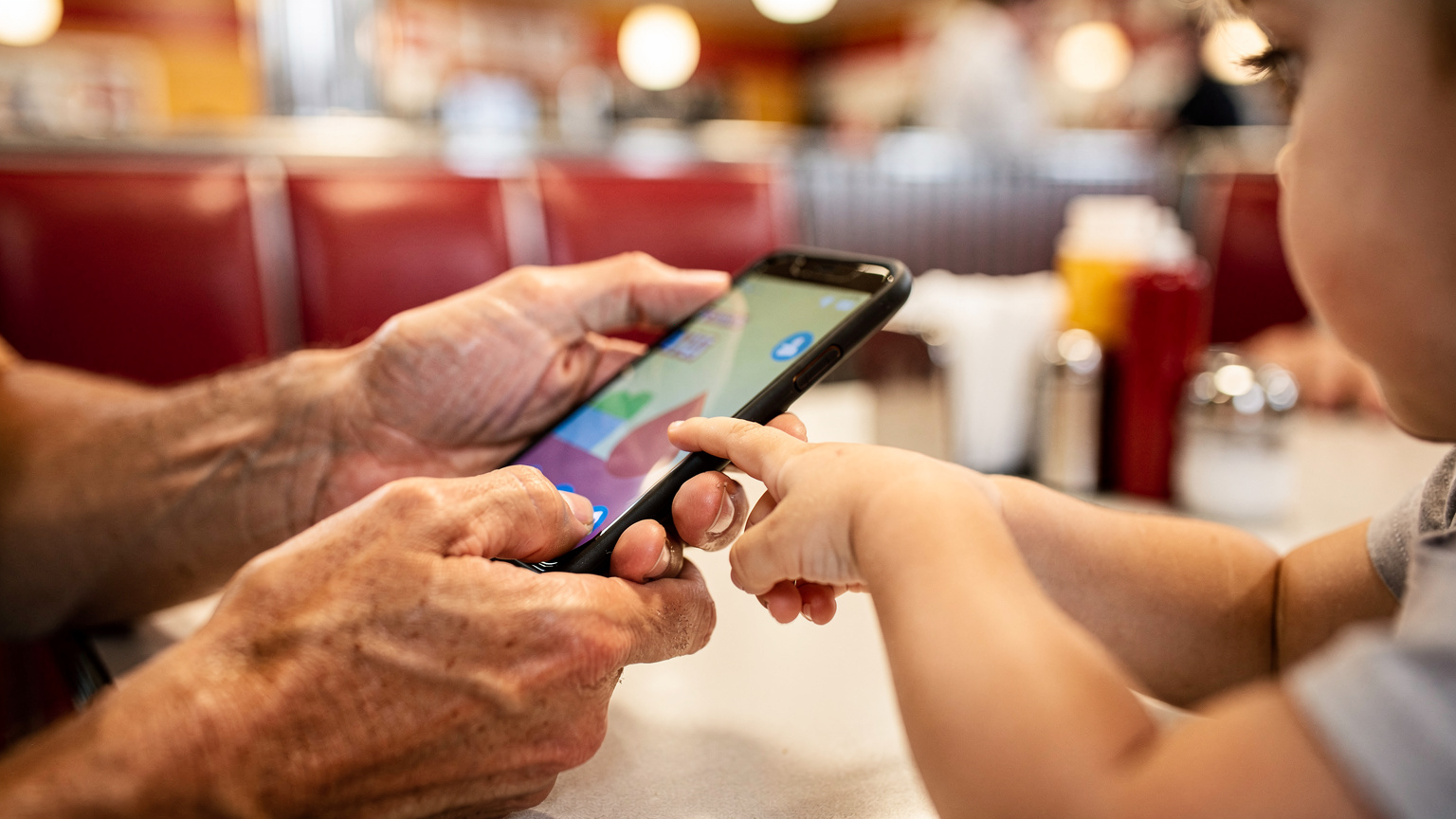A close-up of a parent's hands holding a smartphone, with a child's hand (aged 6-8) reaching to tap the screen, perhaps interacting with an educational app or game on the phone, set against a blurred background of a lively American diner.