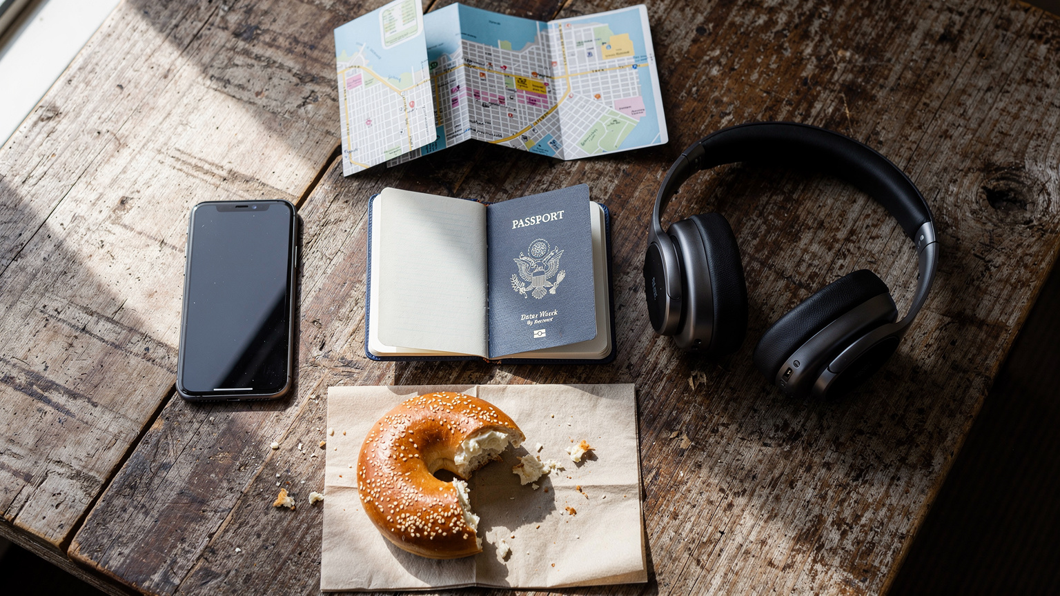 An editorial overhead flat-lay shot on a rustic wooden table, featuring a passport, a smartphone showing a blank or 'no service' screen, a pair of wireless headphones, a small folded paper map of a US city, and a half-eaten bagel with cream cheese, suggesting a traveler's moment of planning or minor frustration.