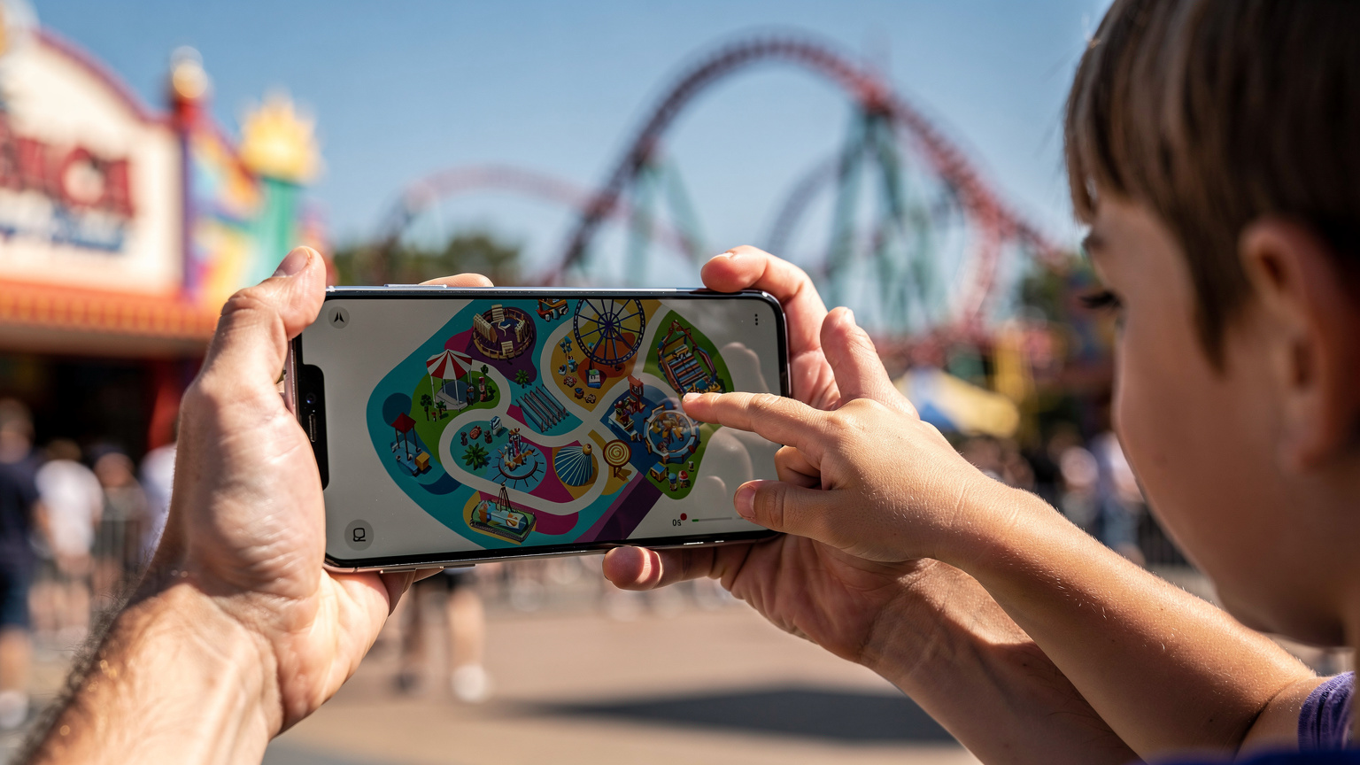 A close-up of a parent's hands, possibly at a theme park, holding a smartphone showing a map of the park, with a child (aged 7-9) eagerly pointing towards a ride in the distance. The background is blurred with colourful rides and crowds.