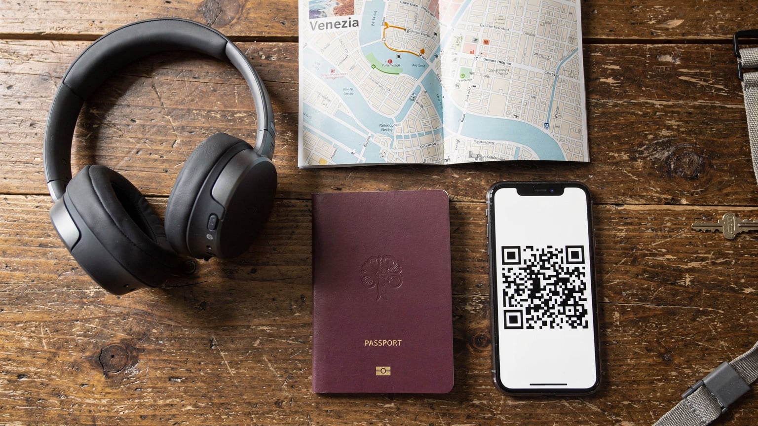 An overhead flat-lay of travel essentials: a passport, a smartphone displaying an eSIM activation QR code, noise-canceling headphones, and a detailed paper map of a European city, all arranged on a rustic wooden table.