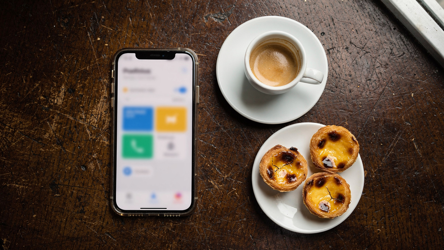 Uma mesa de café em Lisboa com um smartphone mostrando a interface de um aplicativo de operadora móvel, um copo de café e alguns pastéis de nata, indicando uma pausa para trabalho ou planejamento.