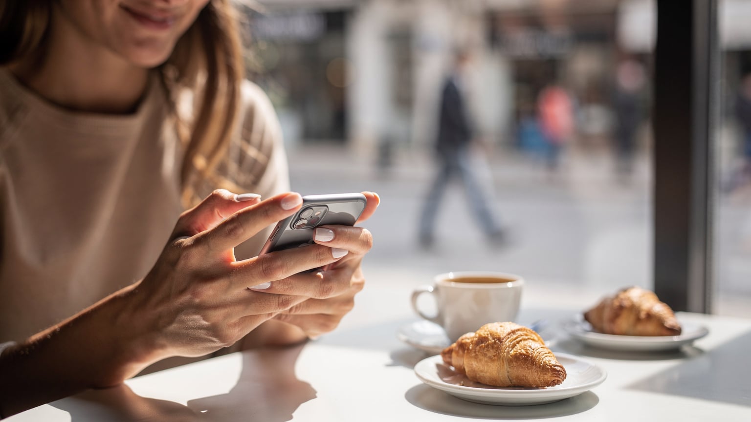 Žena s úsmevom inštaluje eSIM na svojom iPhone, drží ho v ruke pri káve v kaviarni v Tirane, Albánsko, s rozostreným pozadím mestskej ulice.