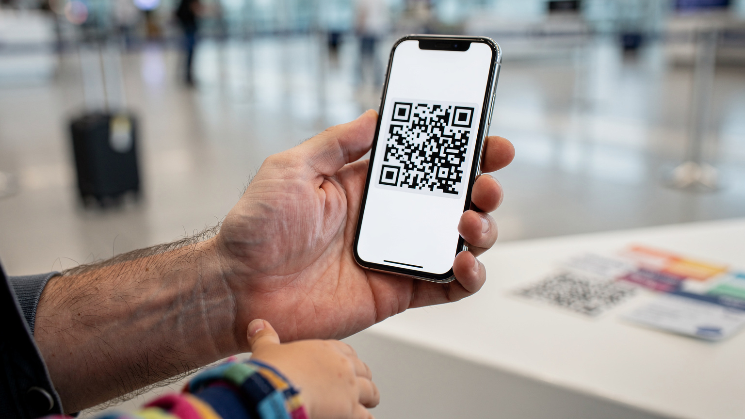 Manos de un padre sosteniendo un smartphone, mostrando un código QR de activación de eSIM, mientras la pequeña mano de un niño mira con curiosidad por encima de su hombro en un mostrador de facturación de aeropuerto, simbolizando la preparación del viaje.