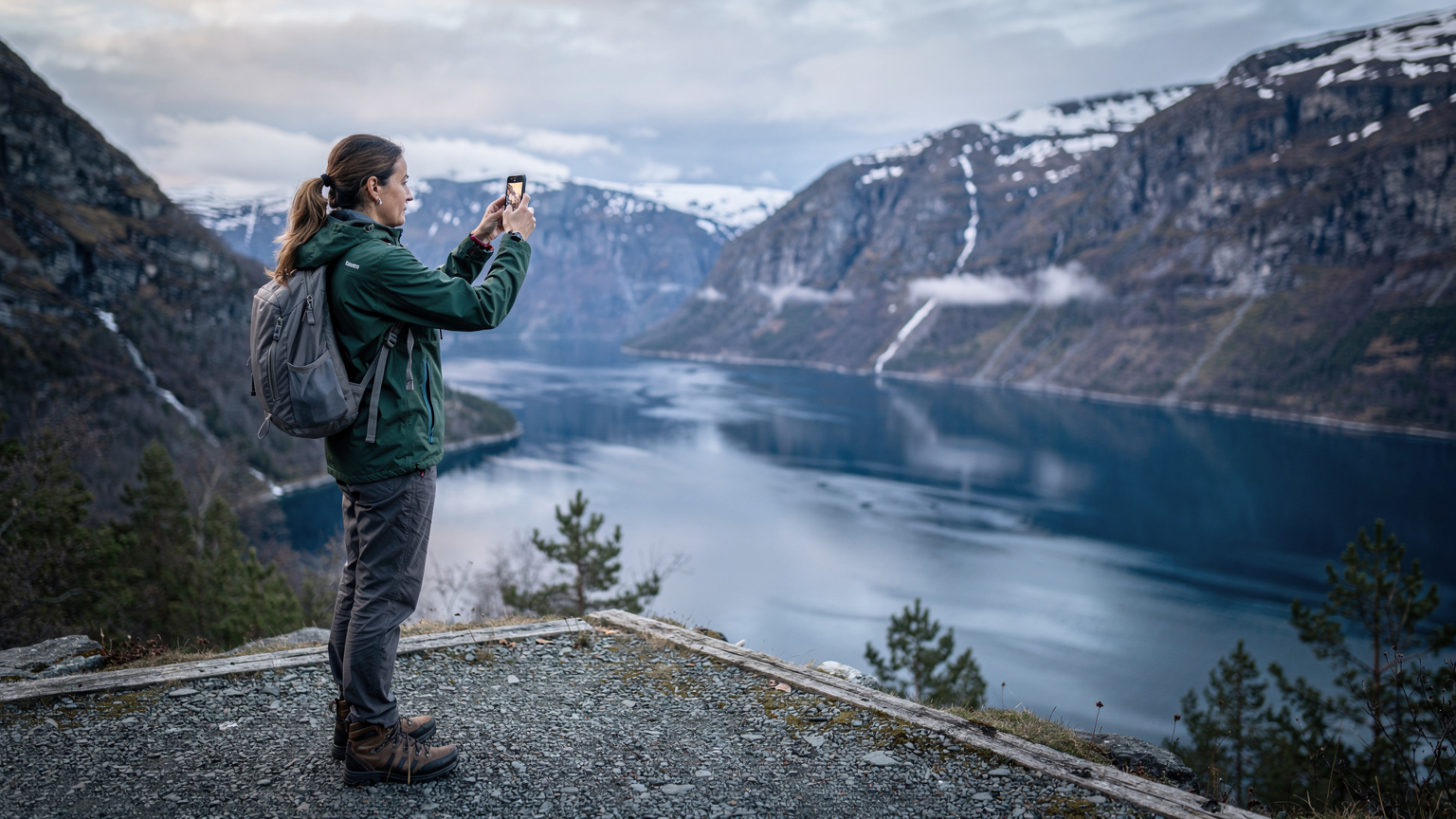 En kvinde holder sin smartphone op for at fange et billede af det dramatiske norske landskab, med en fjord og fjelde i baggrunden, tydeligt optaget i et område med sparsom befolkning.