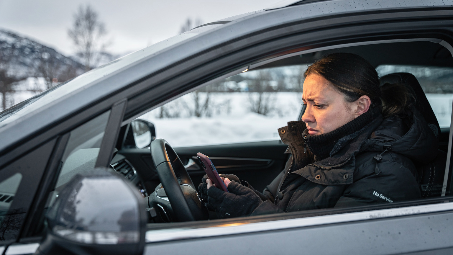 En kvinde rynker panden, mens hun kigger frustreret på sin smartphone, der viser "ingen service", mens hun sidder i en bil parkeret ved et afsidesliggende, snedækket norsk landskab.