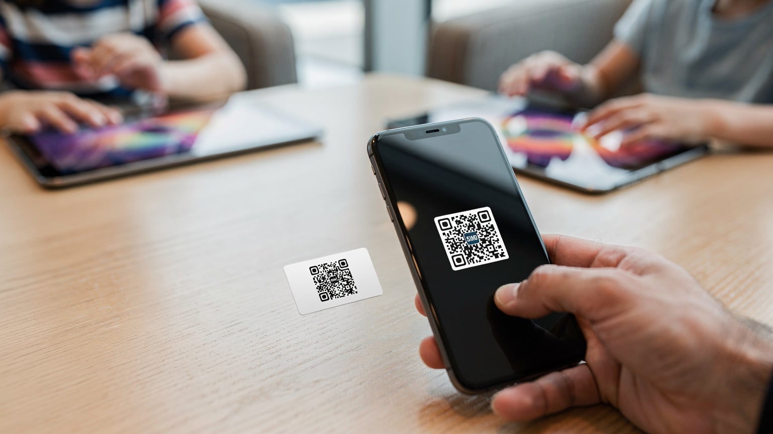 Eine Nahaufnahme eines iPhones, das einen Cellesim-QR-Code scannt, während im unscharfen Hintergrund zwei Kinderhände Tablets halten und darauf spielen, alles auf einem Holztisch in einer Flughafen-Lounge.