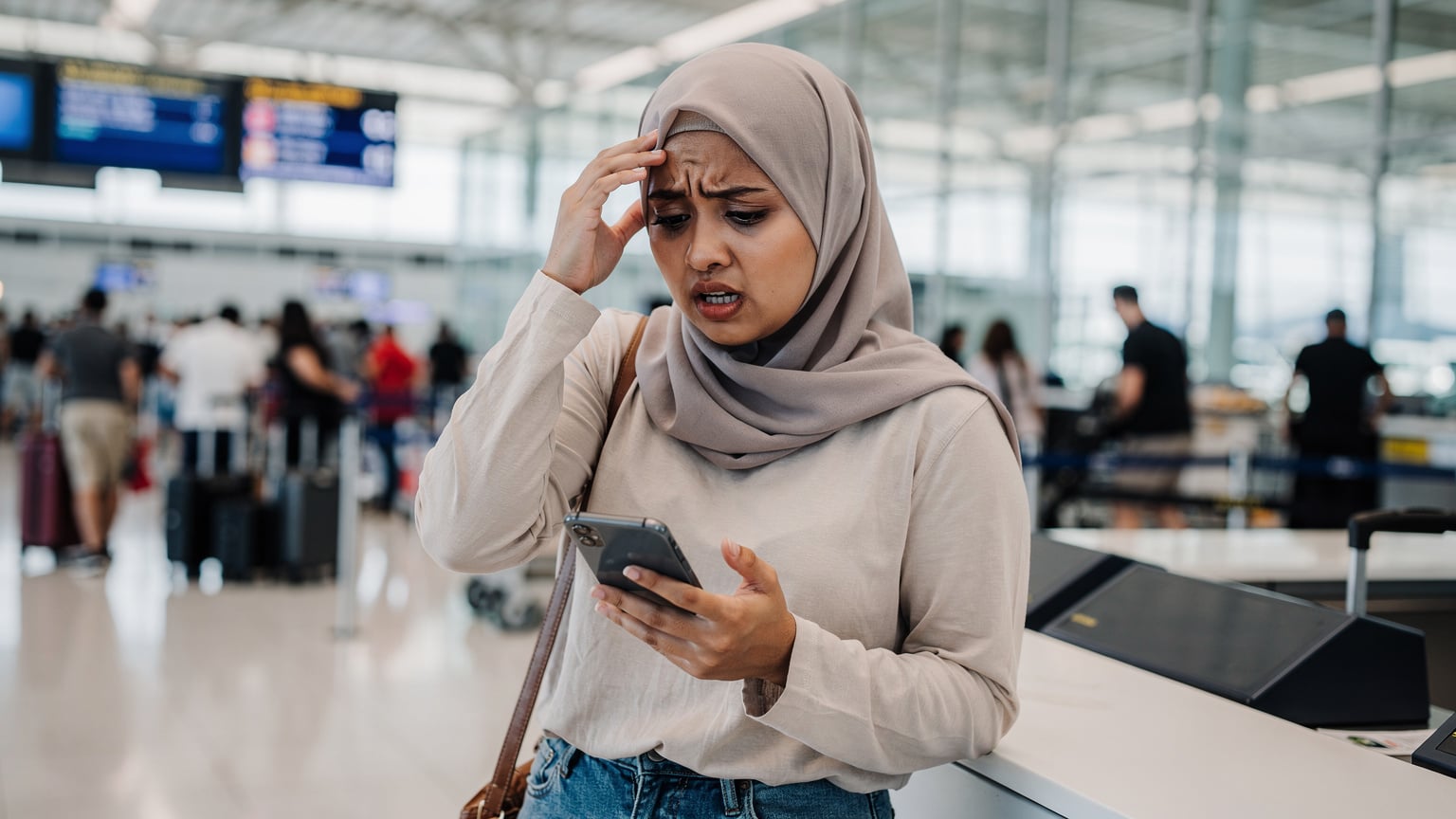 Seorang wanita melihat iPhone dengan riak bimbang, memegang kepalanya di kaunter daftar masuk lapangan terbang yang sibuk, menunjukkan masalah sambungan data.