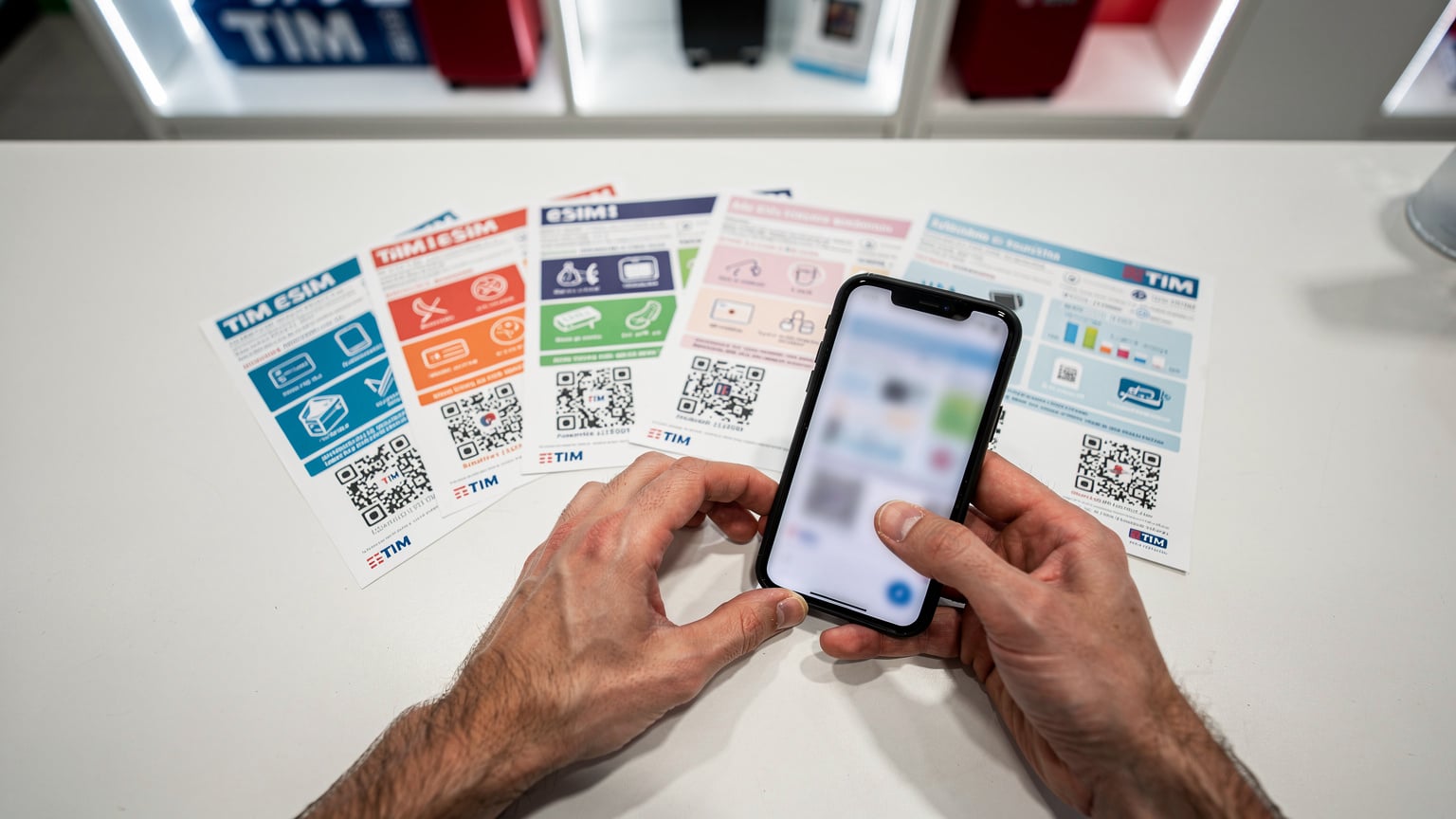 A traveler meticulously compares various TIM eSIM tourist plan brochures and QR codes displayed on a counter at a TIM store in Rome's Termini Station, focusing on data allowances and validity periods.