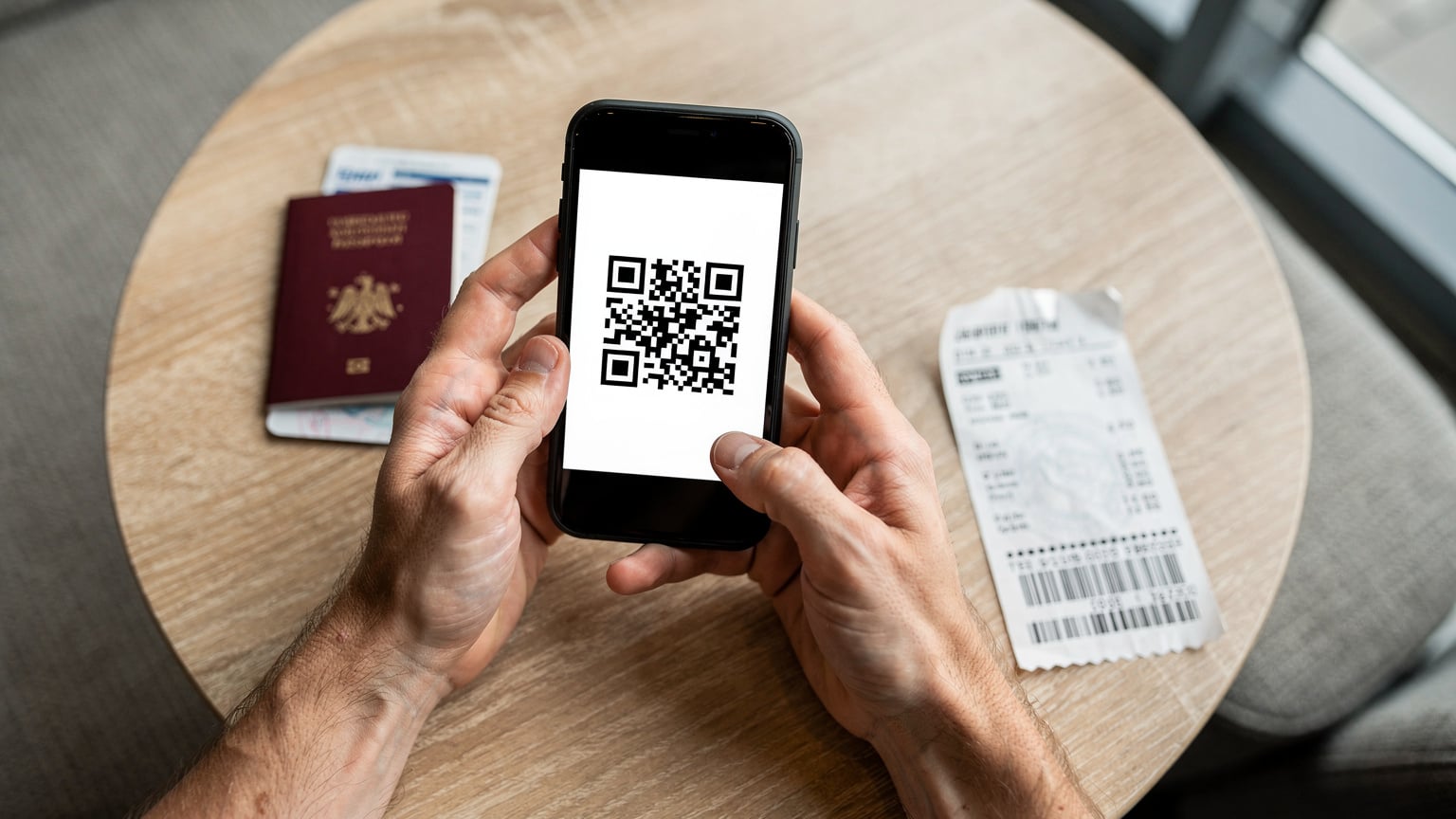 Un viajero escanea un código QR en su smartphone para activar su eSIM, con un pasaporte abierto y una tarjeta de embarque arrugada sobre una mesa de madera clara de una cafetería.