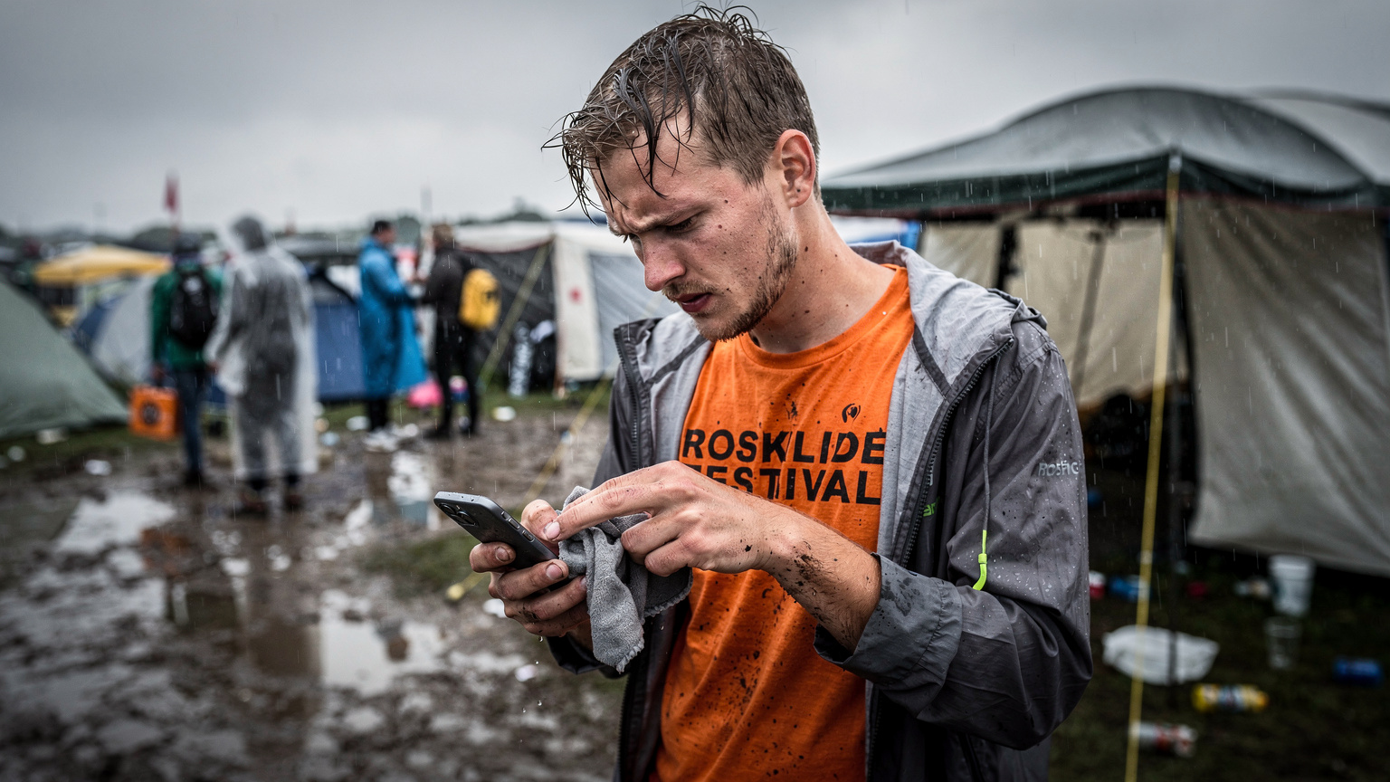 En ung festivalgænger i en regnfuld Roskilde Festival-lejr prøver at tørre sin smartphone af med en klud, mens han ser frustreret på skærmen, der viser manglende signal. Teltduge og mudret jord er synlige i baggrunden.