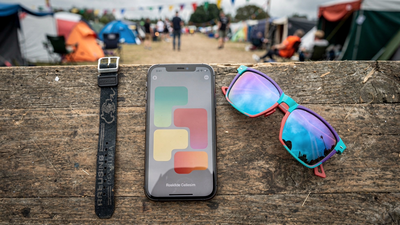 Et festivalarmbånd, en smartphone med Cellesim-appen åben og et par farverige solbriller ligger på en rå træoverflade, med den slørede baggrund af en travl festivalcampingplads.