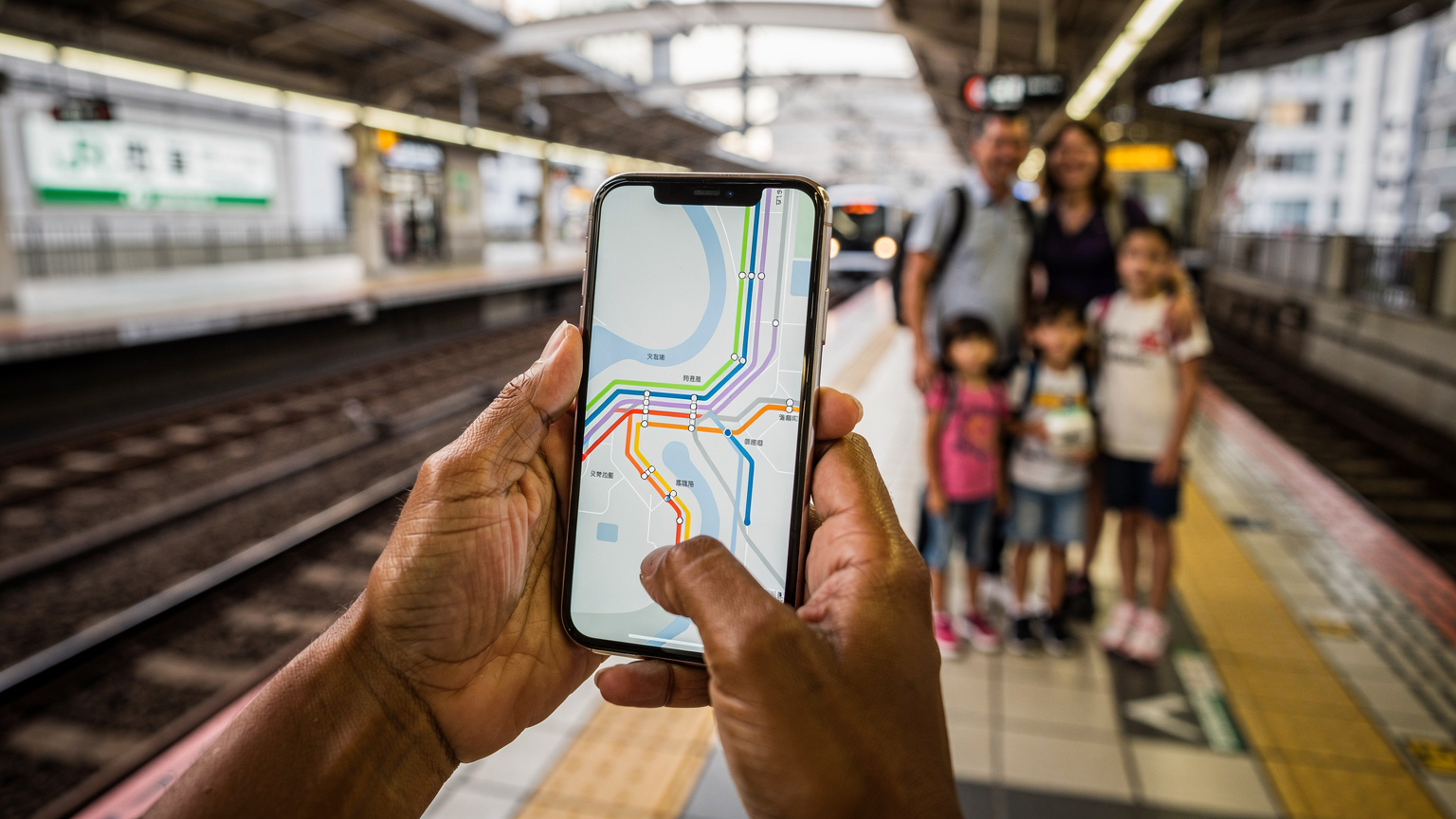 Mãos de uma mulher brasileira segurando um smartphone com o Google Maps aberto, mostrando uma rota de metrô em Tóquio, com a estação Shinjuku ao fundo, enquanto a família (fora de foco) espera em uma plataforma.