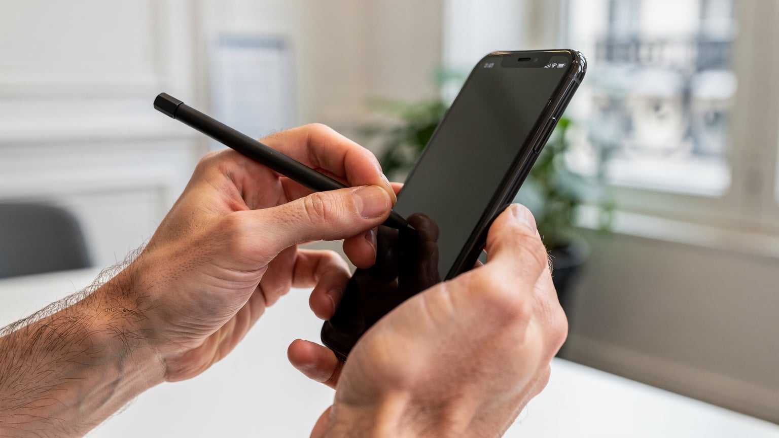 Gros plan sur des mains d'homme tapotant l'écran d'un iPhone avec un stylet, dans un espace de co-working moderne à Paris. L'écran est flou pour éviter les informations sensibles, mais le geste est clair.