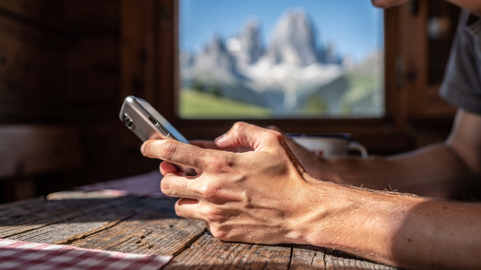 Mani di un giovane viaggiatore che scorrono un'app di messaggistica su uno smartphone moderno, con un vibrante sfondo sfocato di un paesaggio montano italiano, mentre si rilassa su una panchina di legno di un rifugio alpino.
