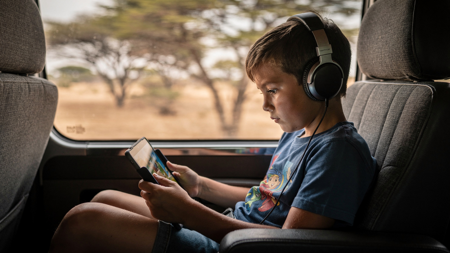 A child, aged about 9, with headphones on, engrossed in a tablet displaying a downloaded animated movie, sitting comfortably in the back seat of a safari transfer vehicle.
