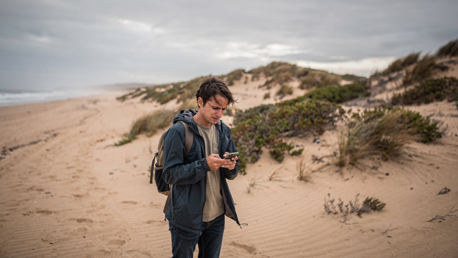 Un viajero consulta su teléfono en una playa remota de la Costa de la Luz, con dunas y vegetación salvaje al fondo, buscando señal con una leve frustración.