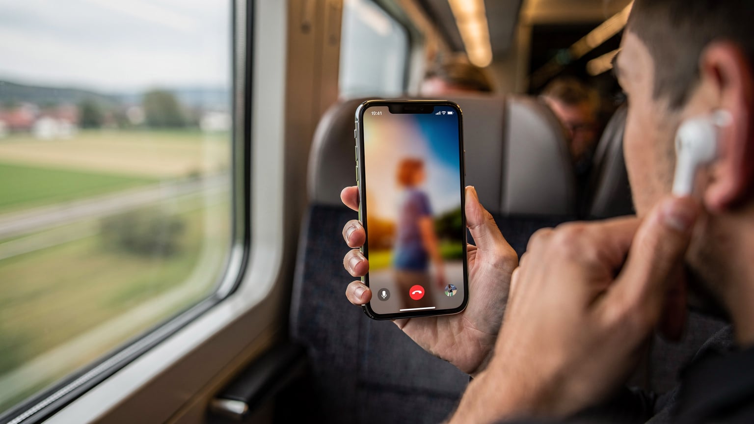 En tæt på-optagelse af en hånd, der holder en iPhone 18 og foretager et videoopkald i et travlt togmiljø, måske på Interrail 2026. Skærmen viser sløret videoindhold, og den anden hånd holder en hovedtelefon tæt på øret. En togvinduet med forbipasserende landskab er sløret i baggrunden.