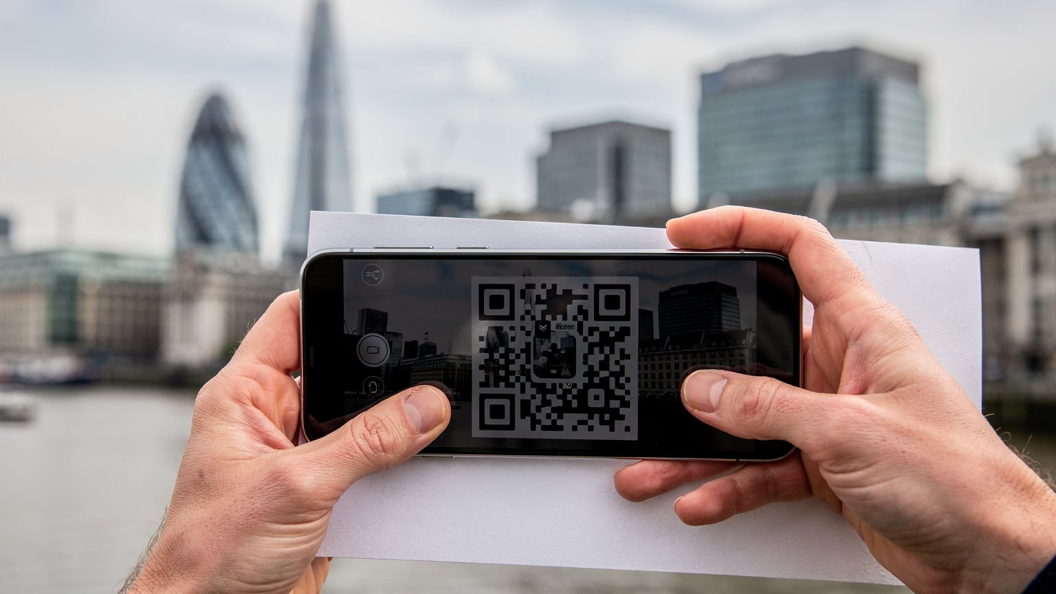 Un primo piano delle mani di una persona che tiene uno smartphone e scansiona un codice QR su un foglio stampato, con lo skyline di Londra sullo sfondo sfocato, suggerendo un'attivazione eSIM in viaggio.