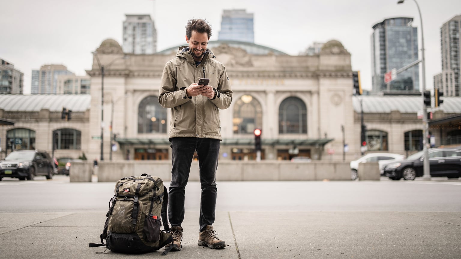Un voyageur souriant, sac à dos à ses pieds, utilisant son téléphone pour consulter une carte numérique devant la gare historique de Via Rail à Vancouver, avec le centre-ville en arrière-plan et un ciel couvert mais lumineux.