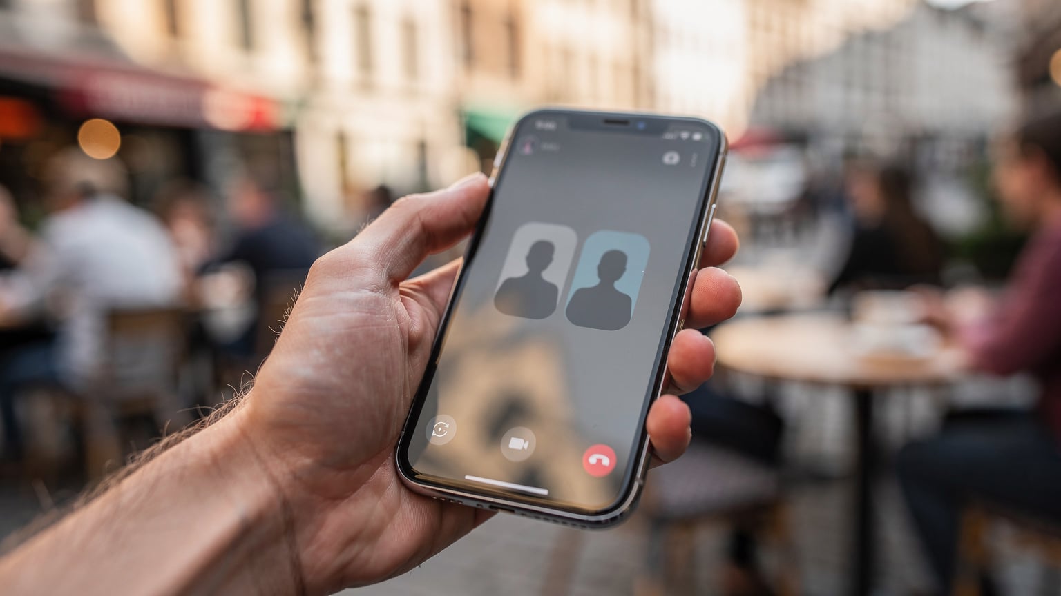 Mână apropiată, ținând un smartphone cu ecranul pe o aplicație de mesagerie, afișând un apel video în desfășurare, cu un fundal estompat dintr-o terasă de cafenea din Bruxelles.