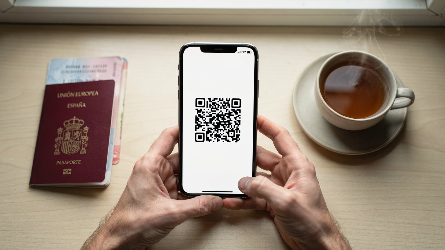 Manos de una persona sosteniendo un smartphone, la pantalla muestra el lector de código QR para la activación de una eSIM. En la misma mesa, un pasaporte español abierto y una taza de té humeante. El fondo es un escritorio de madera claro, con una estética de preparación de viaje.