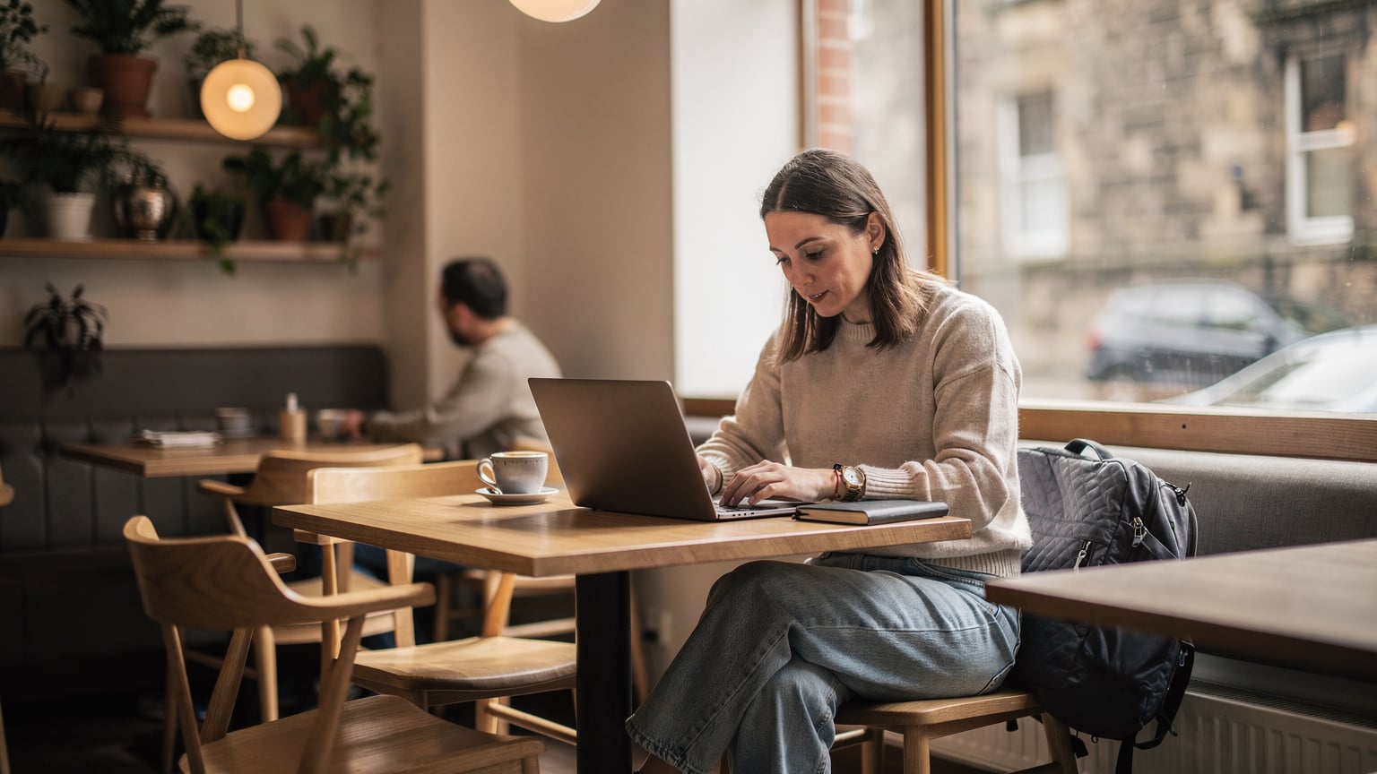 Una mujer con gafas, sentada en una mesa de madera en una cafetería luminosa en el barrio de Leith, Edimburgo, usando su portátil y su teléfono. La mesa tiene una taza de café y un cuaderno. Detrás de ella, se ven estanterías con libros y una ventana que da a la calle. La atmósfera es relajada y productiva.