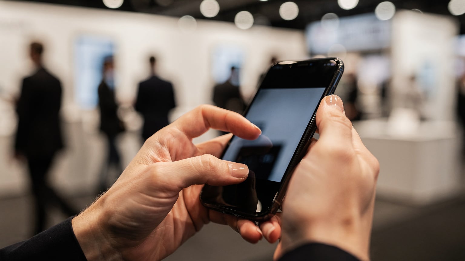 Крупный план женских рук, настраивающих eSIM на экране смартфона, на фоне размытых огней и движения выставочного зала.