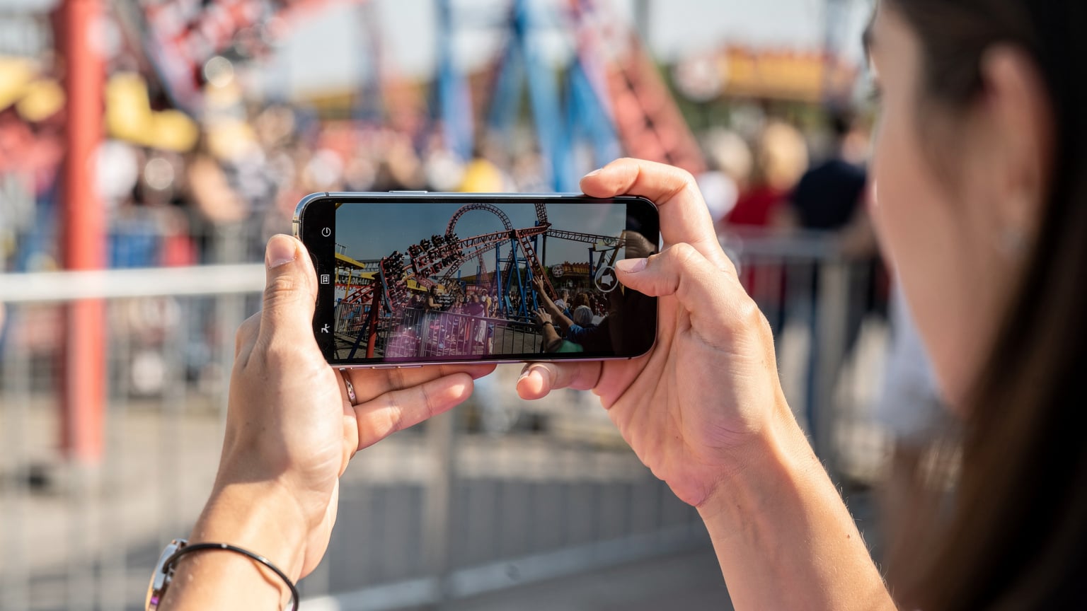 Nahaufnahme einer weiblichen Hand, die ein Smartphone hochhält, um ein Video von lachenden Freunden auf einer Achterbahnfahrt aufzunehmen. Im Vordergrund sind die Hände der Person zu sehen, die ein Zeichen der Begeisterung zeigen.
