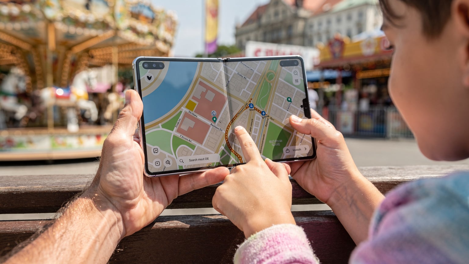 Ruky rodiča a dieťaťa spoločne listujú v offline mape na skladacom telefóne Samsung Galaxy Z Fold 8, zatia ľ čo sedia v zábavnom parku v Budapešti, v pozadí je vidieť kolotoč.